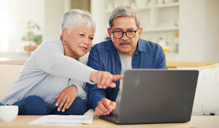 Older couple on a couch looking at a laptop, the woman pointing at the screen as the man watches closely in a bright living room.
