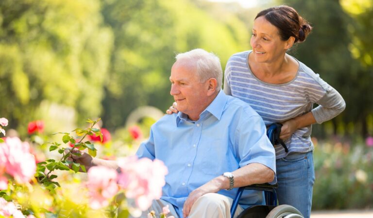 Older man in a light blue shirt seated in a wheelchair in a flower garden, smiling while a woman stands behind him, helping and smiling.
