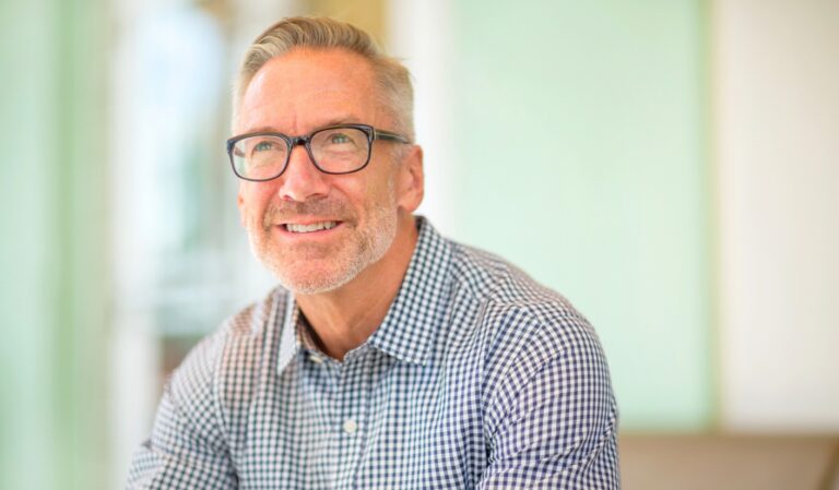 Smiling middle-aged man with gray hair and glasses wearing a blue-and-white checkered shirt, in a bright, softly blurred indoor setting.